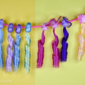 May include: A collection of colorful doll hair extensions hanging from a pink string against a yellow background. The extensions feature various shades of blue, purple, pink, and yellow, with decorative butterfly and flower-shaped clips.