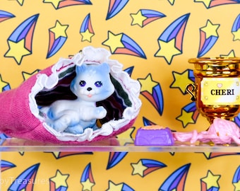 Pode incluir: Um gatinho de brinquedo azul e branco espreita de uma meia rosa com acabamento branco. Um troféu dourado com o nome "CHERI" está ao lado de uma tigela de comida roxa e fio rosa. O fundo é amarelo com formas de estrelas e cometas.