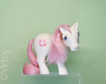 May include: A white toy pony with pink hair and tail, and blue eyes. The pony has a pink floral design on its side. The toy is standing on a clear, rectangular surface against a light green background.
