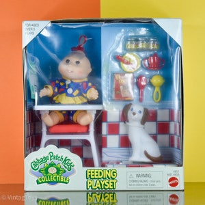 Op de afbeelding: Cabbage Patch Kids voedingsspeelset in originele verpakking. De set bevat een pop in een kleurrijke outfit, een kinderstoel en voedingsaccessoires. Een wit hondenbeeldje is ook inbegrepen. De doos is gelabeld "Voor kinderen vanaf 3 jaar."