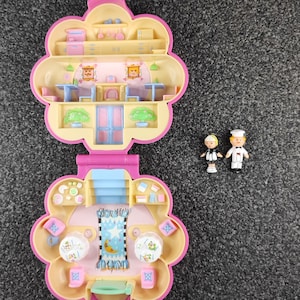 Può includere: Un playset Polly Pocket rosa e giallo vintage, aperto per rivelare due livelli di un edificio in miniatura. Il playset ha la forma di un fiore. Due piccole statuette sono accanto al playset. Uno è uno chef e l'altro una cameriera.
