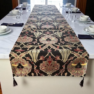 May include: A long, black table runner with an ornate floral pattern in shades of gold, red, and gray. The runner has a tassel at each end and is placed on a white surface. The table runner is approximately 108 inches long.