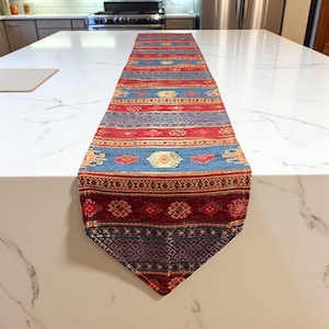 May include: A rectangular table runner with a geometric pattern. The runner features bands of red, blue, and gold. The design includes repeating shapes and patterns. The runner is made of a woven fabric.