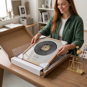Op de afbeelding: Een ingelijste print van een vintage vinylplaat. De plaat is zwart met een gouden label en omringd door tekst. De lijst is van licht hout en de print zit in een kartonnen doos. De print is gesigneerd en gedateerd.