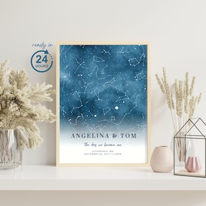 May include: A framed print featuring a blue watercolor background with a constellation design. The print includes the names "ANGELINA & TOM" and the text "The day we became one" with a date and location. The frame is a light beige color.