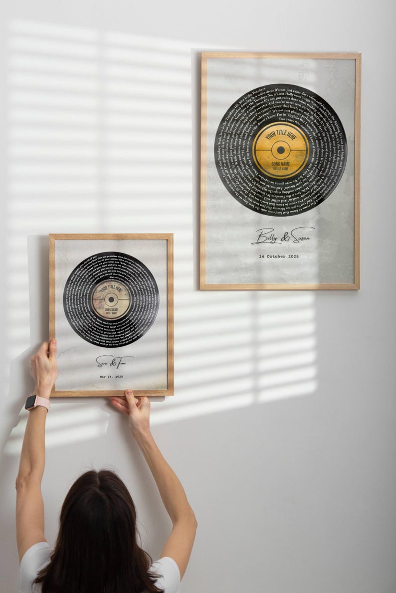 Op de afbeelding: Twee ingelijste kunstdrukken met een vinylplaatontwerp. De platen hebben een zwart oppervlak met witte tekst, een geel middellabel en zijn geplaatst tegen een grijze achtergrond. De frames zijn van licht hout. E&eacute;n print wordt vastgehouden.