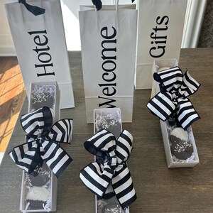 May include: Three white gift bags with black text reading "Hotel", "Welcome", and "Gifts". Each bag has a black bow. Below are boxes with chocolate-covered treats, decorated with black and white striped bows.