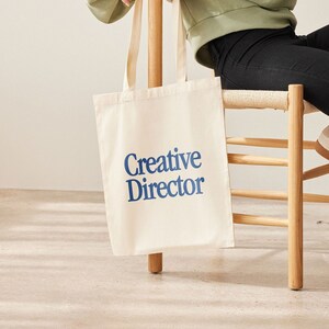 May include: A white canvas tote bag with blue text that reads "Creative Director".