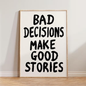 Może przedstawiać: Oprawiony wydruk artystyczny z napisem "BAD DECISIONS MAKE GOOD STORIES" pogrubioną, czarną czcionką. Grafika ma kremowe tło i jest oprawiona w jasne drewno. Wydruk jest prezentowany na drewnianej podłodze.