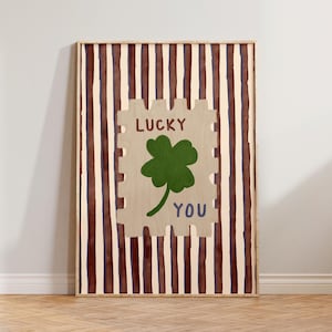 May include: A framed art print featuring a beige stamp design with a green four-leaf clover and the words "LUCKY YOU" in brown and blue. The background has vertical brown and beige stripes. The artwork is displayed on a wooden floor.