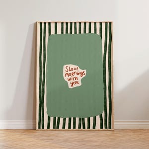 Pode incluir: Uma impressão artística emoldurada com uma borda listrada verde e branca. A obra de arte apresenta uma grande forma verde com o texto "Slow mornings with you" em vermelho. A moldura é de cor castanho claro.