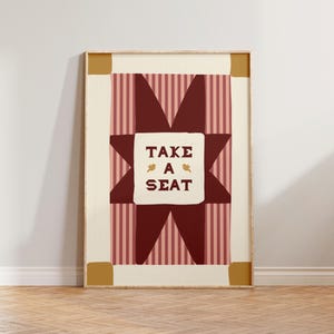 May include: A rectangular wall art print in a wooden frame featuring a central white square with the text "TAKE A SEAT" in dark red, flanked by two small gold birds. The background consists of a dark red star shape over pink vertical stripes.