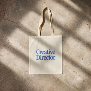 May include: A white canvas tote bag with blue text that reads "Creative Director".
