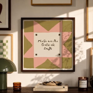 May include: A square framed wall art piece featuring a central white square with the text "These are the Good old Days" in dark brown, surrounded by a pink star shape on an olive green background, with four small dark brown star icons.