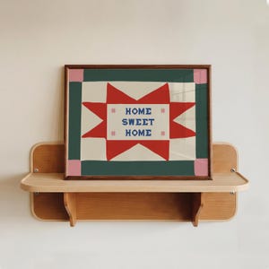 Puede incluir: Una impresi&oacute;n rectangular enmarcada que presenta una forma de estrella roja sobre un fondo crema con el texto azul HOME SWEET HOME y cuatro peque&ntilde;os acentos de estrellas rosas. El marco es de madera marr&oacute;n oscura y el borde es verde oscuro con esquinas rosas.