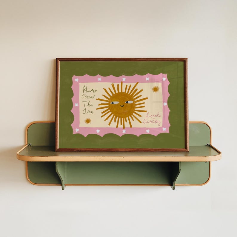 Puede incluir: Una impresi&oacute;n art&iacute;stica enmarcada con una ilustraci&oacute;n de un sol sonriente y el texto "Here Comes The Sun Little Darling". La obra de arte tiene un borde festoneado rosa y un fondo verde. El marco es marr&oacute;n.