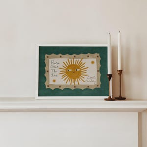 Puede incluir: Una impresi&oacute;n art&iacute;stica enmarcada con una ilustraci&oacute;n de un sol sonriente y el texto "Here Comes The Sun Little Darling". La obra de arte tiene un marco blanco, un borde verde festoneado y se muestra con dos velas blancas en candelabros de lat&oacute;n.