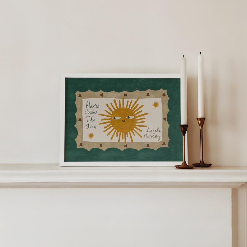 Puede incluir: Una impresi&oacute;n art&iacute;stica enmarcada con una ilustraci&oacute;n de un sol sonriente y el texto "Here Comes The Sun Little Darling". La obra de arte tiene un marco blanco, un borde verde festoneado y se muestra con dos velas blancas en candelabros de lat&oacute;n.