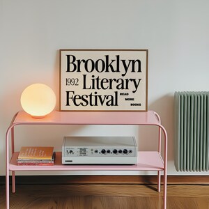 May include: A pale pink metal side table with a spherical lamp and a framed poster reading "Brooklyn 1992 Literary Festival." Beneath, a silver audio device and books are displayed.