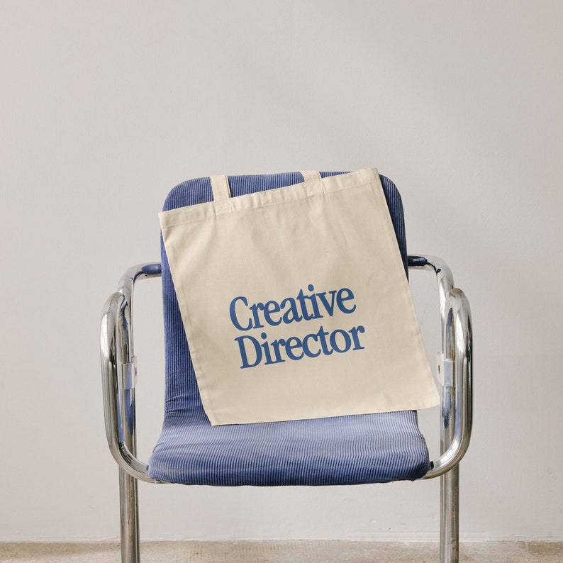 May include: A beige canvas tote bag with blue text that reads "Creative Director" on a blue chair.