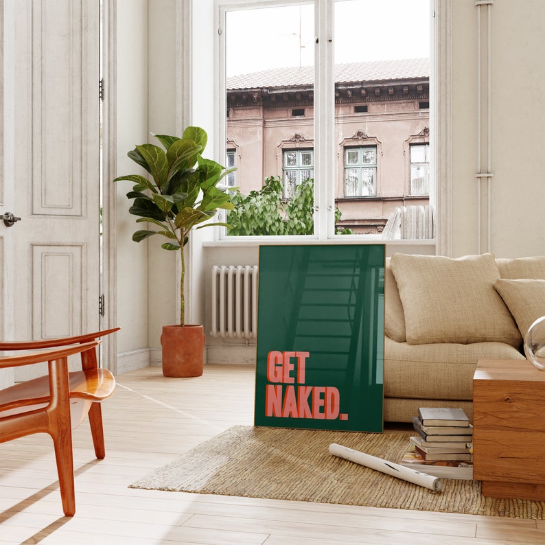 May include: A rectangular framed wall art piece featuring a dark green background with the text GET NAKED in bold, coral-colored capital letters. The artwork is positioned on a woven rug, leaning against a beige sofa in a bright, sunlit room.