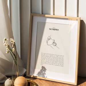 May include: A framed print with a white background and black text that reads "She is Scorpio". The print features a line drawing of a woman's face with a large earring. The print is sitting on a wooden surface with a gold tray and a vase of white flowers.
