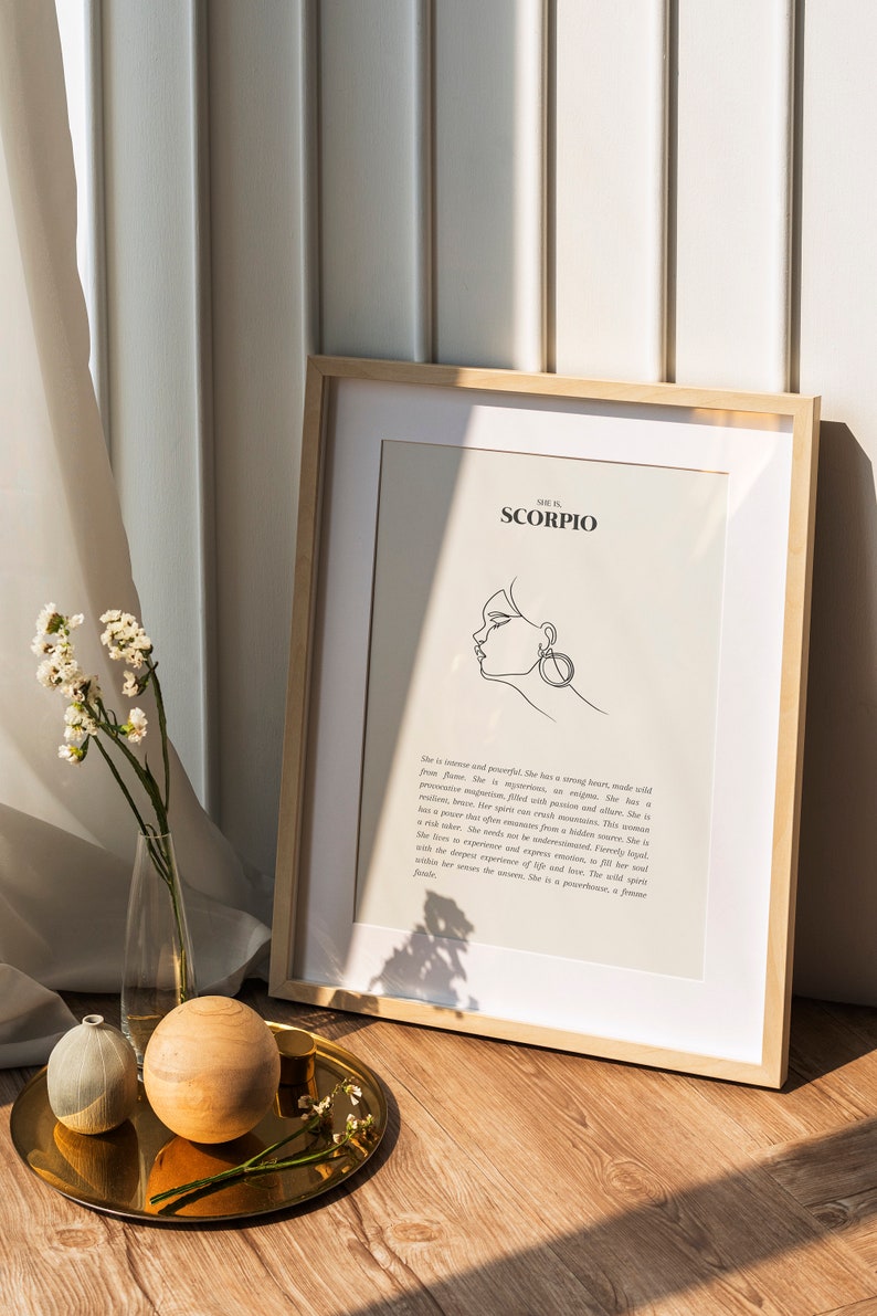 May include: A framed print with a white background and black text that reads "She is Scorpio". The print features a line drawing of a woman's face with a large earring. The print is sitting on a wooden surface with a gold tray and a vase of white flowers.