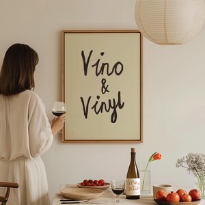 Puede incluir: Una obra de arte enmarcada con las palabras "Vino & Vinyl" en escritura negra cuelga de una pared. Una persona sostiene una copa de vino tinto. Una botella de vino, un plato de tomates y un cuenco de manzanas est&aacute;n sobre una mesa.