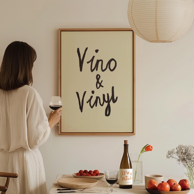 Puede incluir: Una obra de arte enmarcada con las palabras "Vino & Vinyl" en escritura negra cuelga de una pared. Una persona sostiene una copa de vino tinto. Una botella de vino, un plato de tomates y un cuenco de manzanas est&aacute;n sobre una mesa.