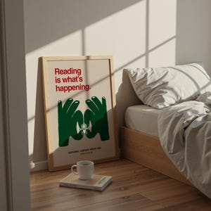 May include: A framed poster with the text "Reading is what's happening" in red, and green hands forming a book shape. Below, it reads "NATIONAL LIBRARY WEEK 1985 APRIL 14 TO 20". A white mug sits on a book, next to the poster. The poster is leaning against a wall.
