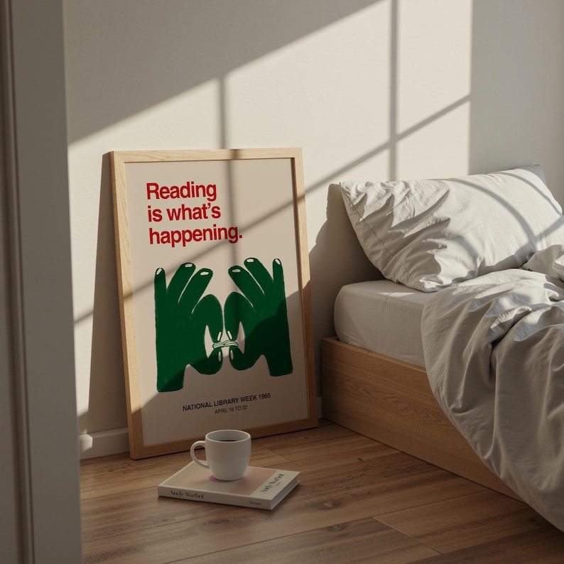 May include: A framed poster with the text "Reading is what's happening" in red, and green hands forming a book shape. Below, it reads "NATIONAL LIBRARY WEEK 1985 APRIL 14 TO 20". A white mug sits on a book, next to the poster. The poster is leaning against a wall.