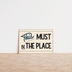 May include: A framed print with a white background and black text that reads "This must be the place". The text is in a stylized font with a blue outline.