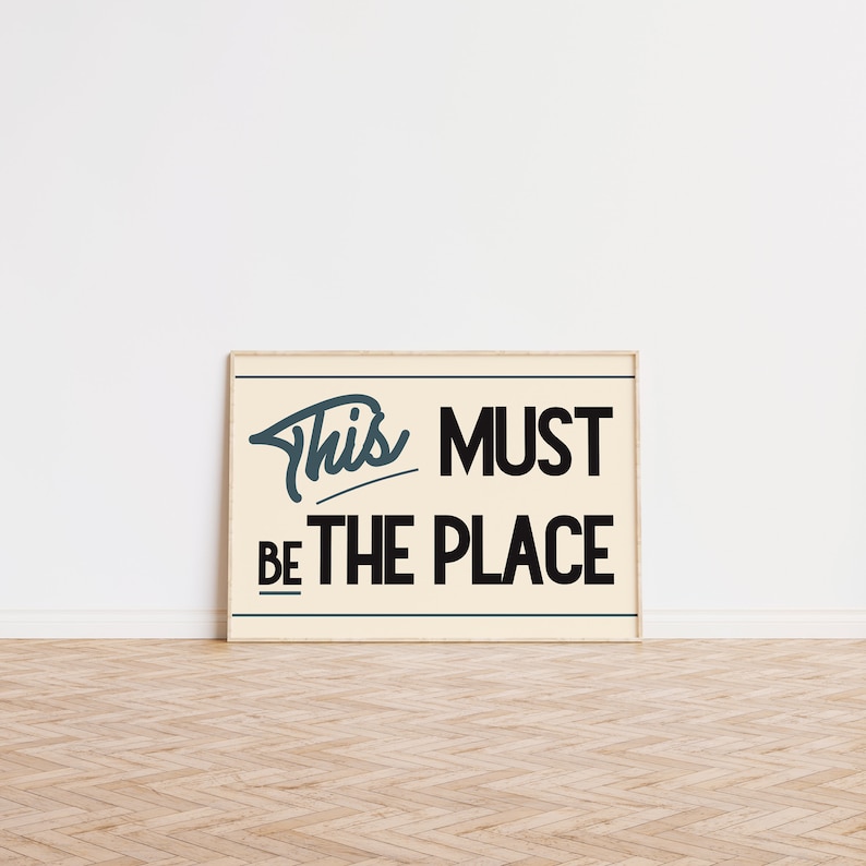 May include: A framed print with a white background and black text that reads "This must be the place". The text is in a stylized font with a blue outline.