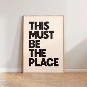 Puede incluir: Una impresi&oacute;n enmarcada con el texto "THIS MUST BE THE PLACE" en letras negras y en negrita sobre un fondo crema.