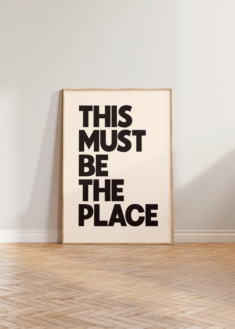Puede incluir: Una impresi&oacute;n enmarcada con el texto "THIS MUST BE THE PLACE" en letras negras y en negrita sobre un fondo crema.