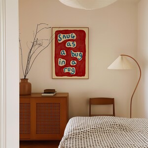 May include: A framed art print with the text "Snug as a bug in a rug" in blue and white on a red background. The artwork hangs on a cream-colored wall above a wooden cabinet and a bed with a checkered blanket. A floor lamp and a vase with branches are also visible.