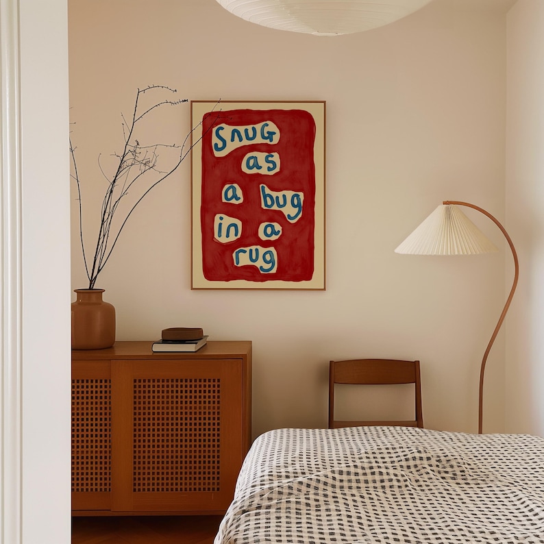 May include: A framed art print with the text "Snug as a bug in a rug" in blue and white on a red background. The artwork hangs on a cream-colored wall above a wooden cabinet and a bed with a checkered blanket. A floor lamp and a vase with branches are also visible.