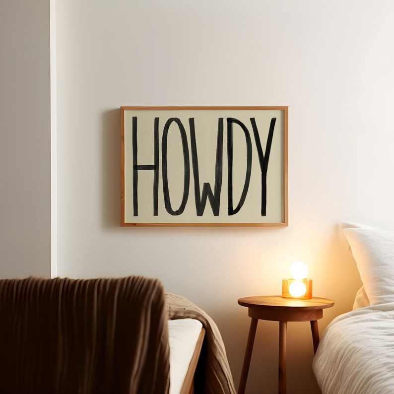 Puede incluir: Una impresi&oacute;n enmarcada con la palabra "HOWDY" escrita en negro sobre un fondo crema. La impresi&oacute;n est&aacute; colgada en una pared blanca sobre una mesita de noche de madera con una l&aacute;mpara.