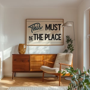 May include: A mid-century modern living room with a wooden cabinet, a beige armchair, a floor lamp, and a framed print with the text "This Must Be The Place".