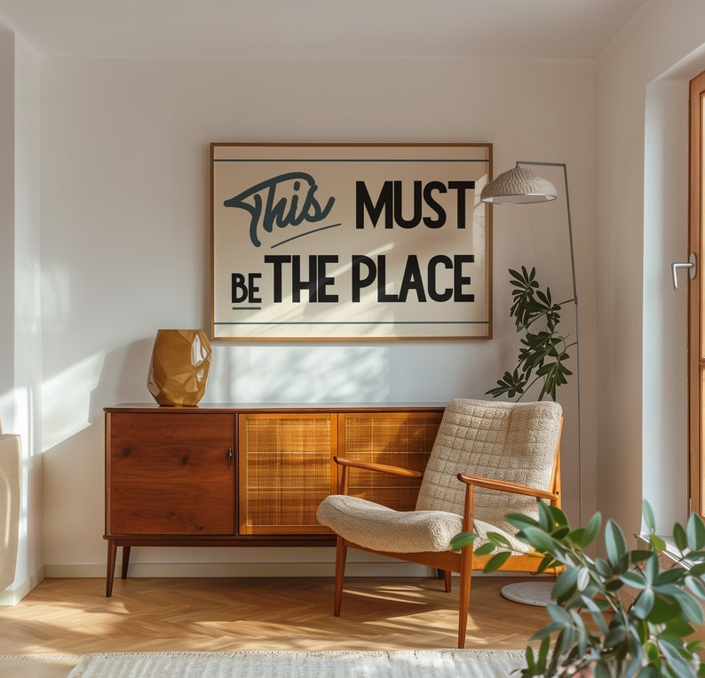 May include: A mid-century modern living room with a wooden cabinet, a beige armchair, a floor lamp, and a framed print with the text "This Must Be The Place".