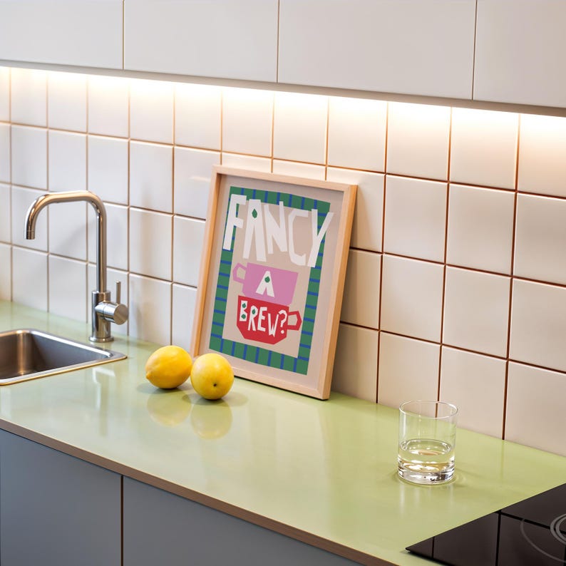 Op de afbeelding: Een ingelijst print met de tekst "Fancy a Brew?" in een kleurrijk, blokkerig lettertype. De print hangt aan een witte betegelde muur boven een groen aanrecht met twee citroenen en een glas water.