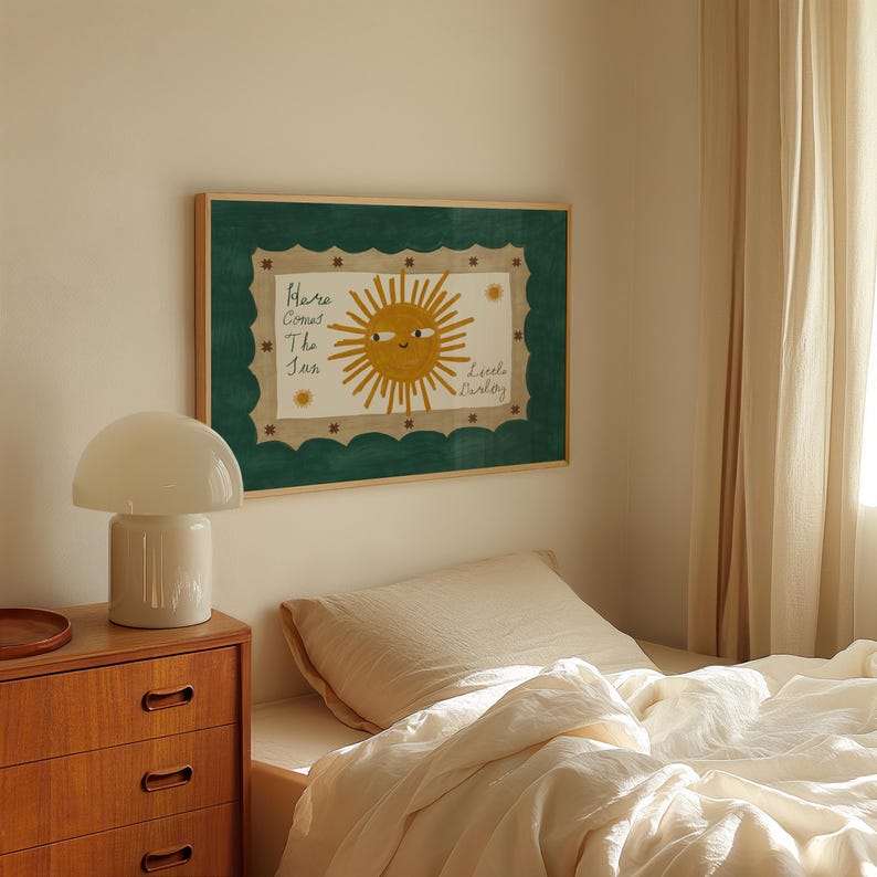 Puede incluir: Una obra de arte enmarcada con una ilustraci&oacute;n de un sol sonriente con el texto "Here Comes The Sun" y "Little Darling". La obra tiene un borde verde festoneado y se muestra sobre una cama con ropa de cama color crema. Una l&aacute;mpara blanca con forma de hongo est&aacute; sobre una c&oacute;moda de madera.