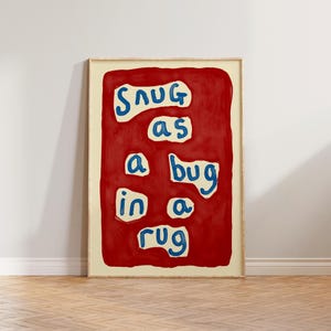 May include: A framed art print with the phrase "Snug as a bug in a rug" in blue letters on a red background. The artwork is in a light wood frame and is displayed against a white wall and wooden floor.