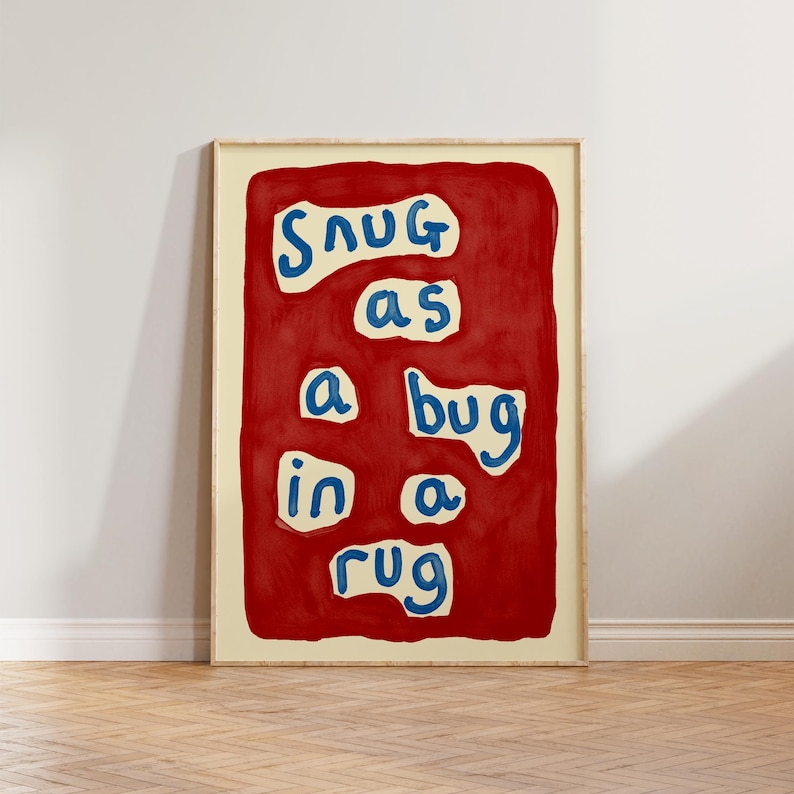 May include: A framed art print with the phrase "Snug as a bug in a rug" in blue letters on a red background. The artwork is in a light wood frame and is displayed against a white wall and wooden floor.