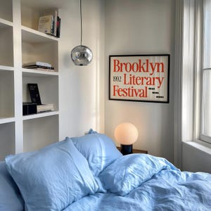 May include: A bedroom interior with a bed dressed in light blue bedding and pillows. A framed poster on the wall displays "Brooklyn Literary Festival 1992." A spherical lamp rests on a bedside table. A chrome pendant light hangs from the ceiling.