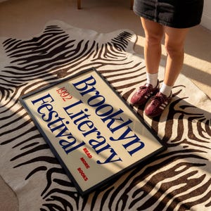 May include: A framed poster with the text "Brooklyn Literary Festival 1992" in blue and red. The poster is on a zebra-print rug. The poster is in a black frame.