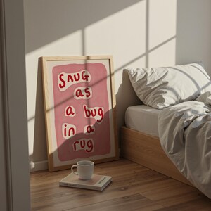 May include: A framed art print with the words "Snug as a bug in a rug" in red lettering on a pink background. The print is leaning against a wall next to a bed with white bedding. A white mug sits on a book on the floor.