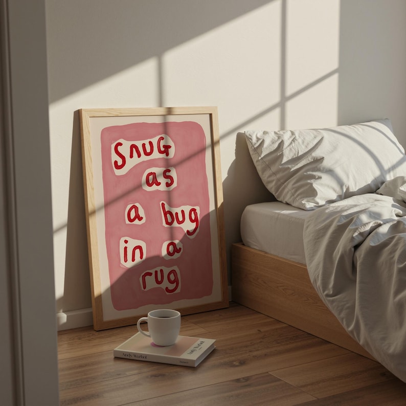 May include: A framed art print with the words "Snug as a bug in a rug" in red lettering on a pink background. The print is leaning against a wall next to a bed with white bedding. A white mug sits on a book on the floor.