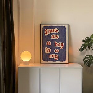 May include: A white cabinet with a framed art print that reads "Snug as a bug in a rug" in red text on a blue background. A round, white table lamp with a glowing yellow orb sits on the cabinet.