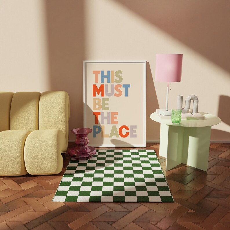 Puede incluir: Una alfombra a cuadros verde y blanca delante de un sof&aacute; amarillo. Una impresi&oacute;n enmarcada con el texto "THIS MUST BE THE PLACE" en letras de colores est&aacute; en la pared detr&aacute;s del sof&aacute;. Una l&aacute;mpara rosa con un vaso verde sobre una mesa auxiliar verde claro.